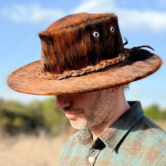 A person wearing a brown cowhide cowboy hat with black brindle patterns, featuring eyelets on the crown.