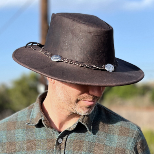 A man wearing a black leather full brim hat with a textured design, featuring a woven rope and charms, outdoors with power lines in the background.