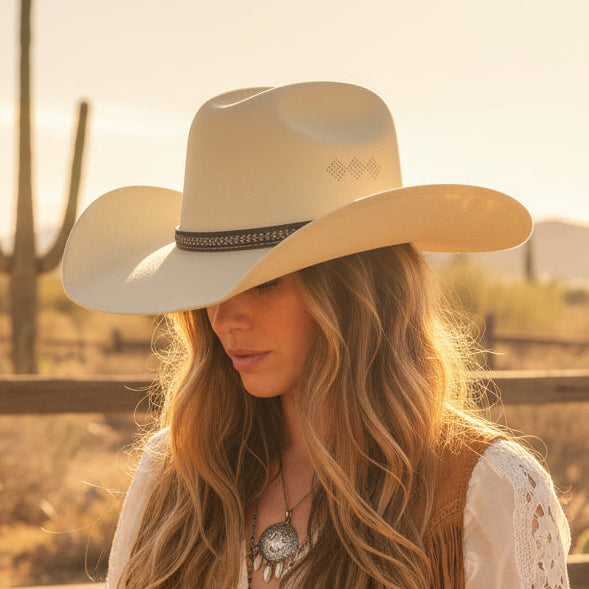 woman wearing a beige cowboy hat with a black band, standing outdoors with a clear blue sky and buildings in the background.